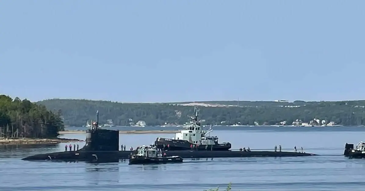 USS New Jersey (SSN-796) on the surface in Halifax Harbour with crew topside, escorted by four Canadian Forces Auxiliary Vessel tugs, with a wooded shoreline and houses behind.