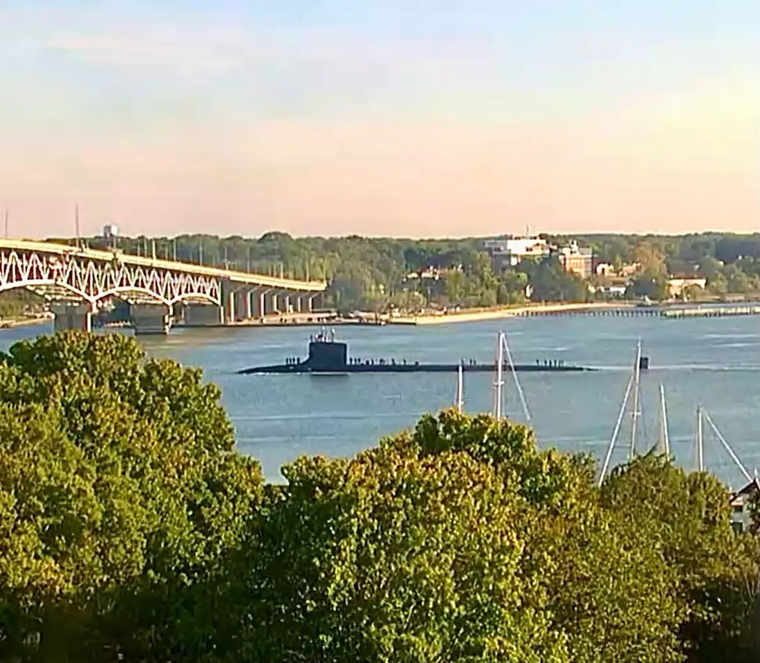 USS New Jersey (SSN-796) on the surface in the York River with crew topside, a steel truss bridge at left and a wooded shoreline behind, seen past treetops and sailboat masts in the foreground.